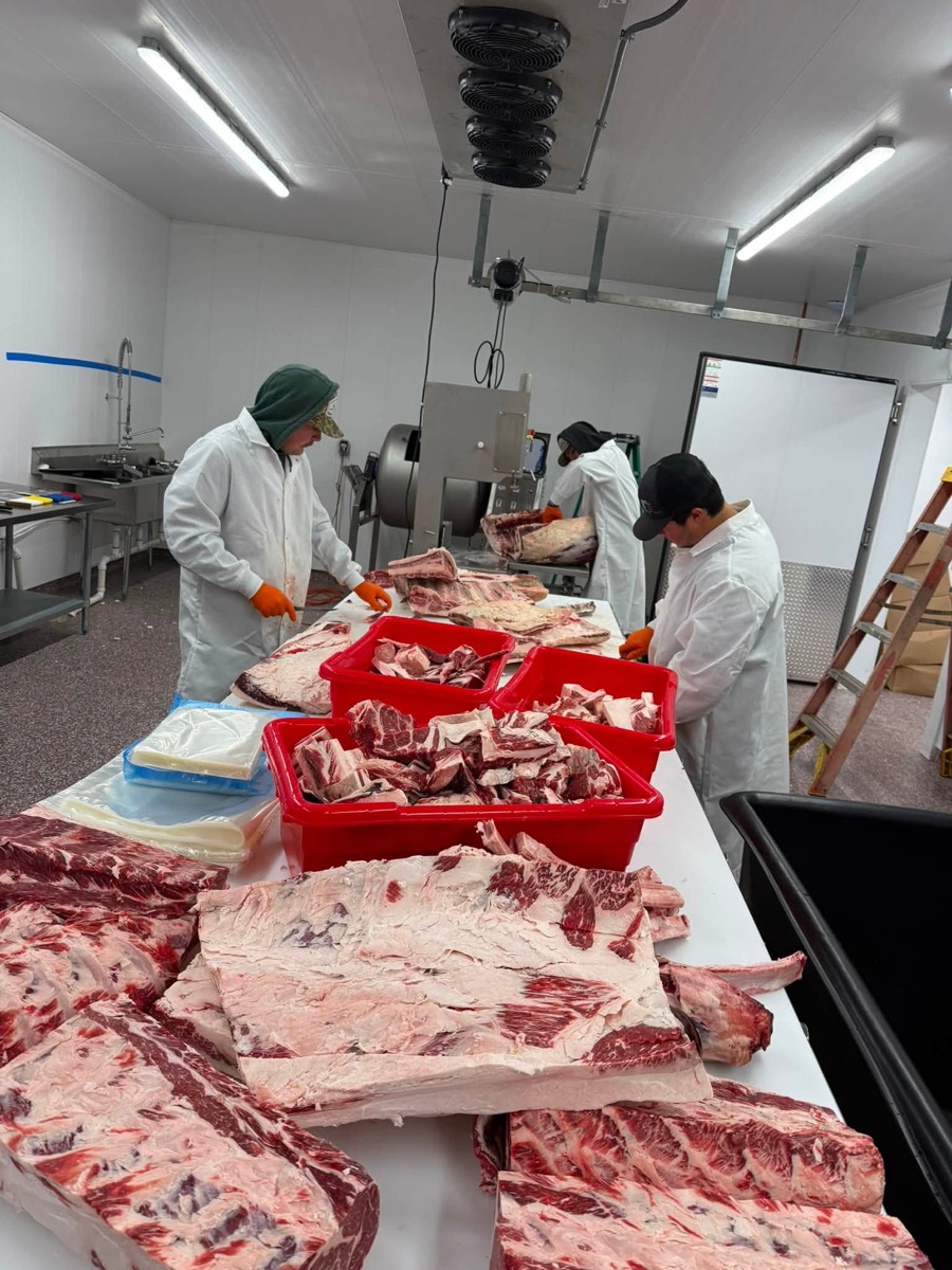 Three butchers breaking down beef at a long cutting table, with red tubs of trimmed cuts.